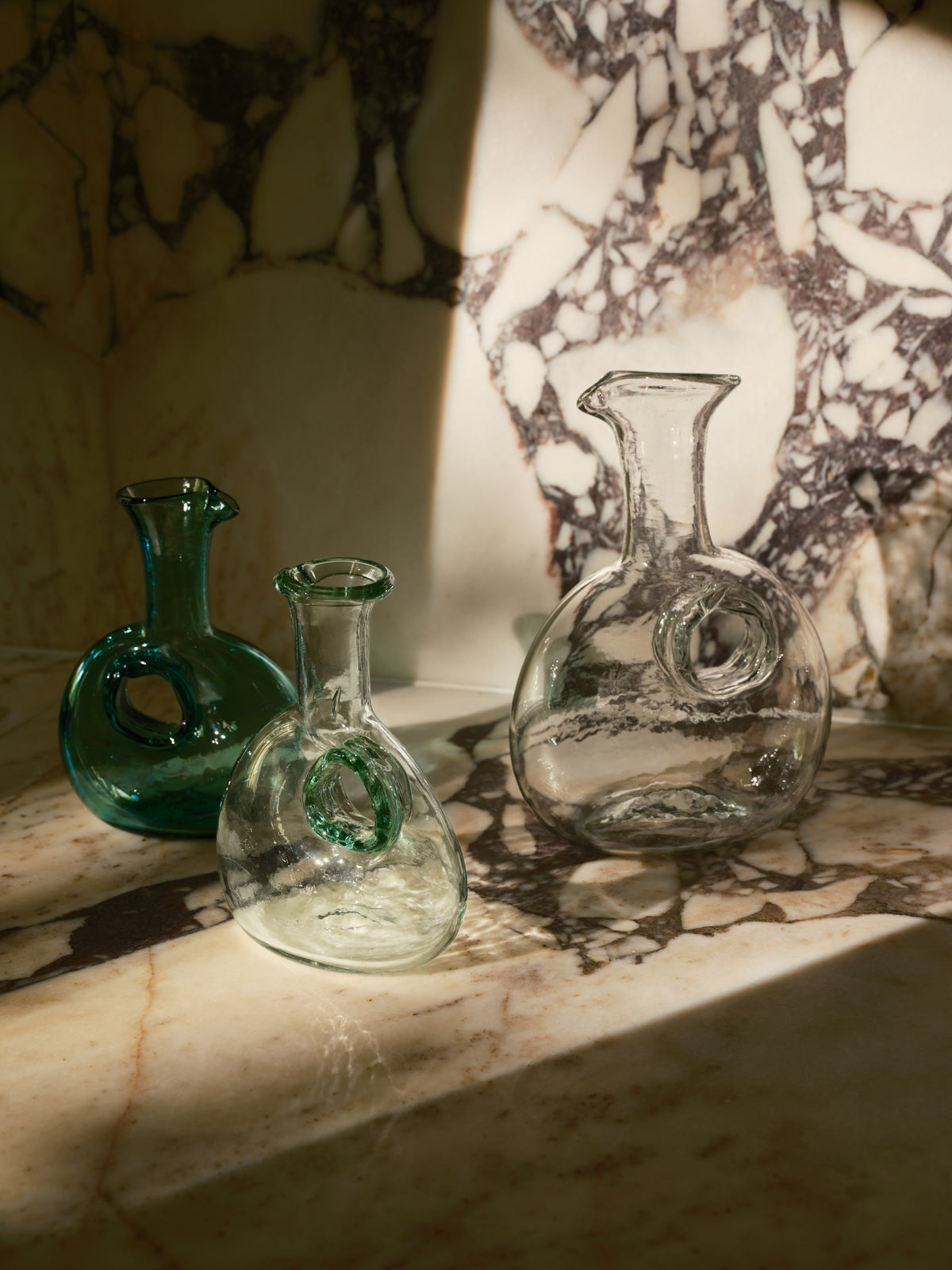 Three decorative glass decanters with looped necks and bulbous bodies, displayed on a marble surface with dramatic sunlight and shadow.