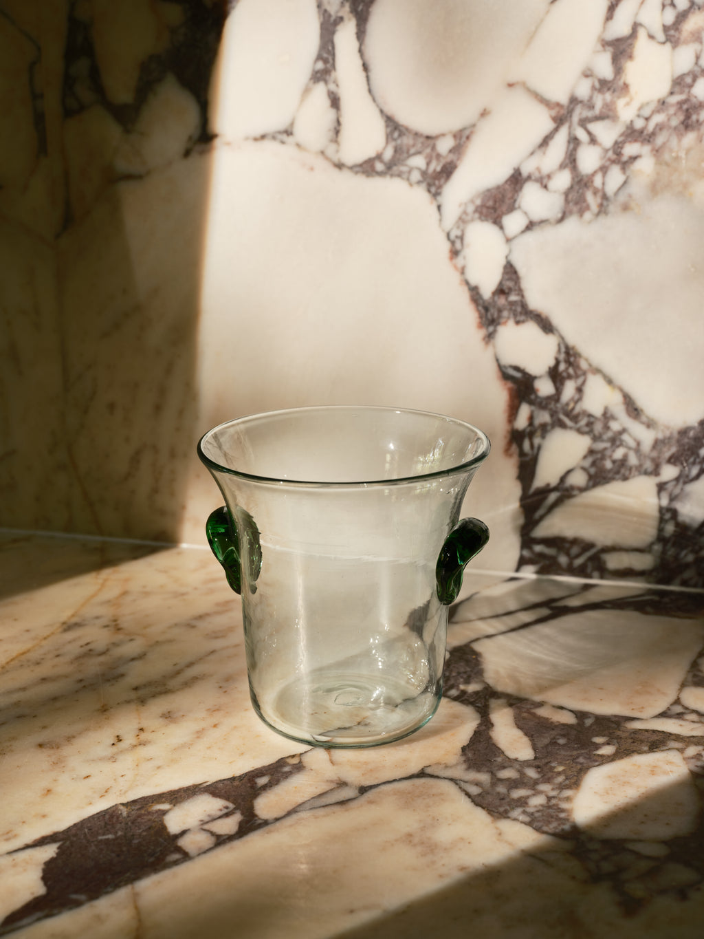 A clear glass cup with two small dark green handles, set on a marble surface against a marble wall with dramatic sunlight and shadows.