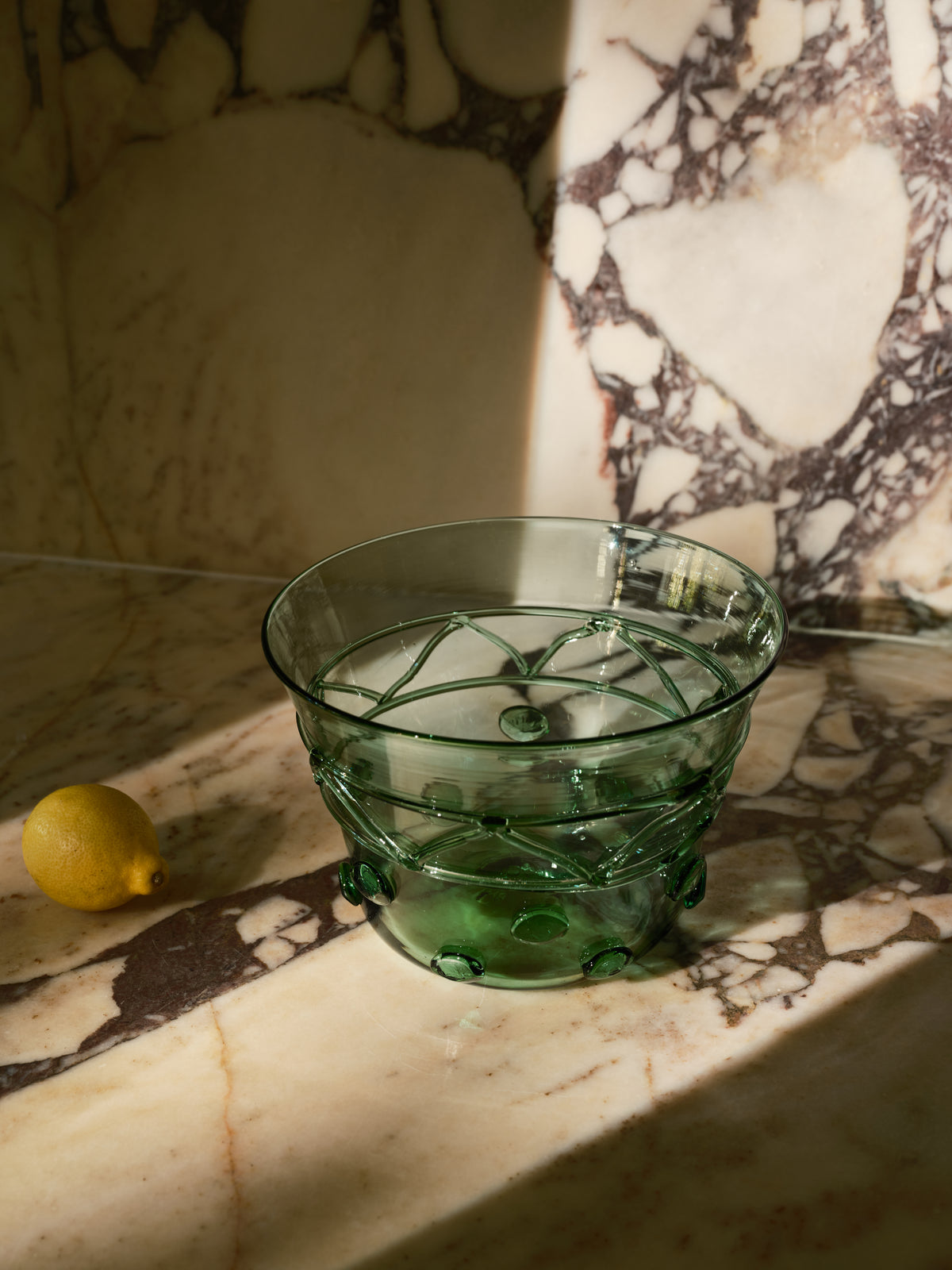 A green transparent glass bowl with a geometric pattern, set on a marble surface with a lemon nearby and dramatic sunlight casting shadows.