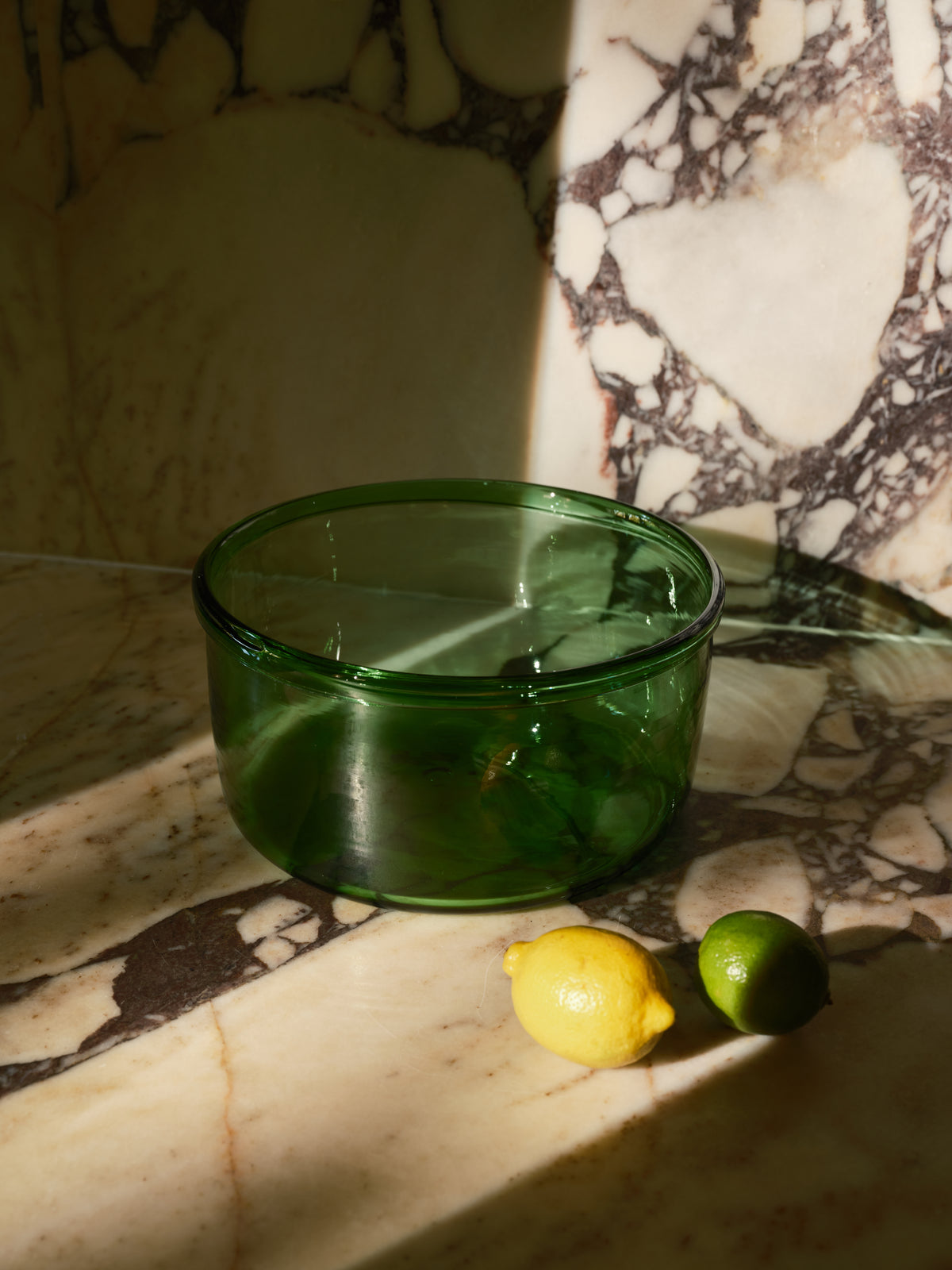 A round translucent dark green glass bowl sits on a marble surface with a lemon and lime beside it, lit by dramatic natural sunlight and shadow.