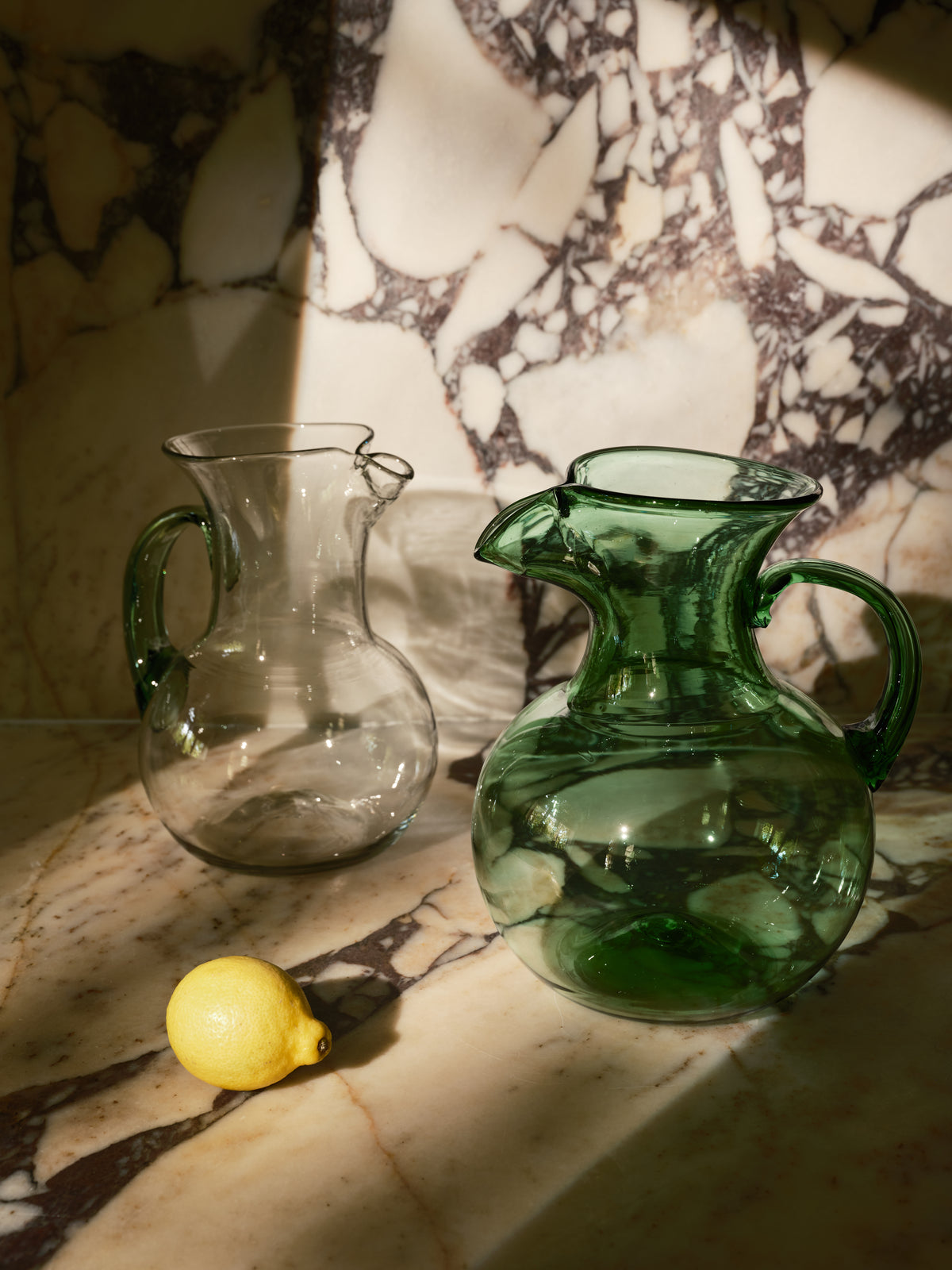 Two glass pitchers on a marble surface with dramatic sunlight and shadow. One pitcher is transparent clear glass, and the other is translucent dark green glass.