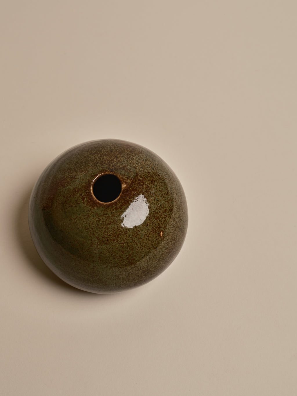A small round ceramic vase with a single opening, featuring a glossy dark green and brown speckled glaze with a white patch, set against a solid beige background.