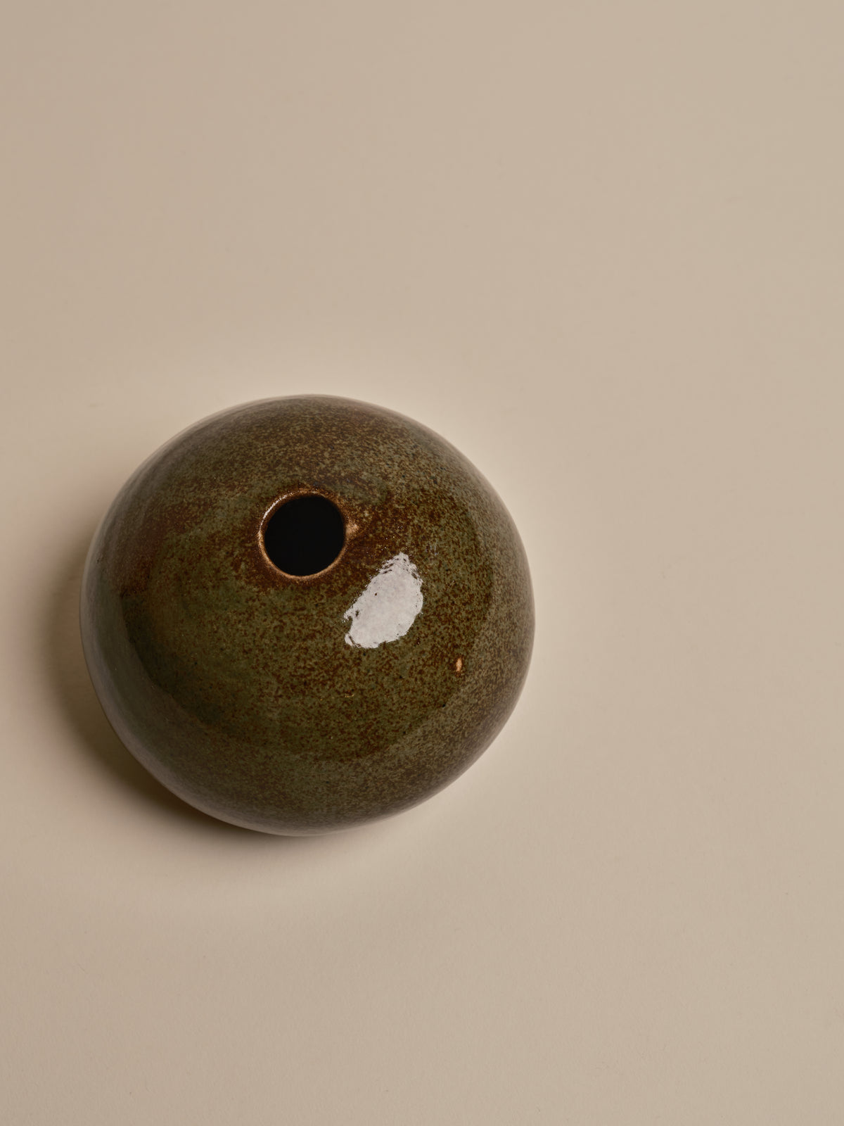 A small round ceramic vase with a single opening, featuring a glossy dark green and brown speckled glaze with a white patch, set against a solid beige background.